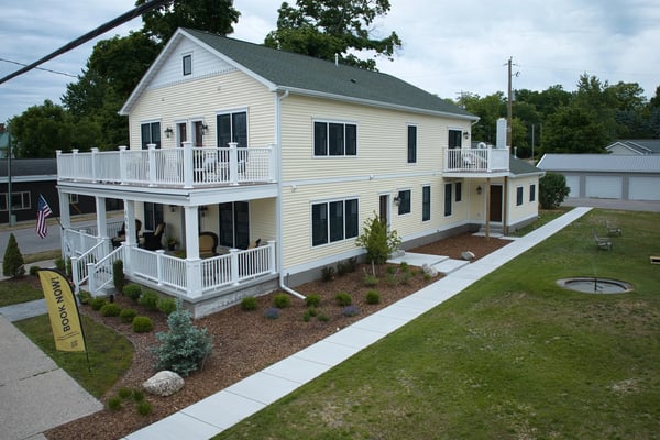 Two-story cream-colored house with white railings, multiple decks, and landscaped front yard with lawn and shrubs.