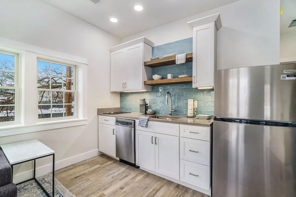 Modern kitchen with white cabinets, stainless steel appliances, blue tile backsplash, wood shelving, and window overlooking trees.